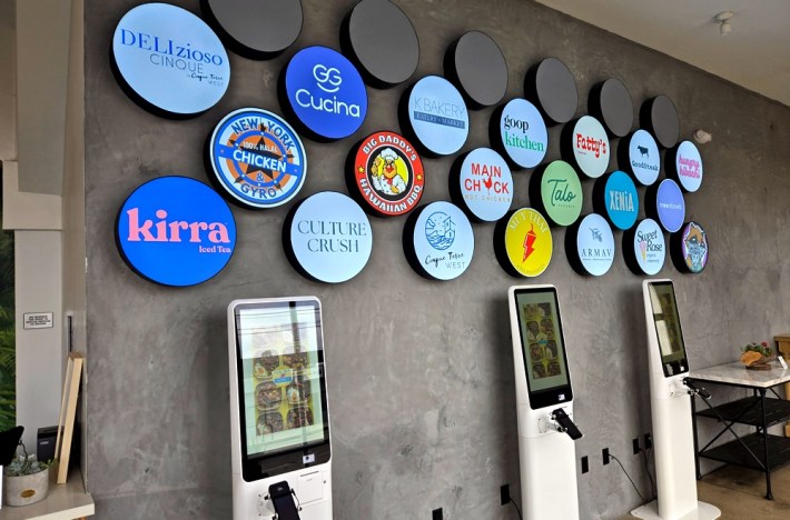 Numerous lit-up circles showing restaurant business name at The Colony in Venice, with self-ordering kiosks below them. Concepts include Main Chick, Kirra Iced Tea, Culture Crush, Talo, Goop Kitchen, and GG Cucina.